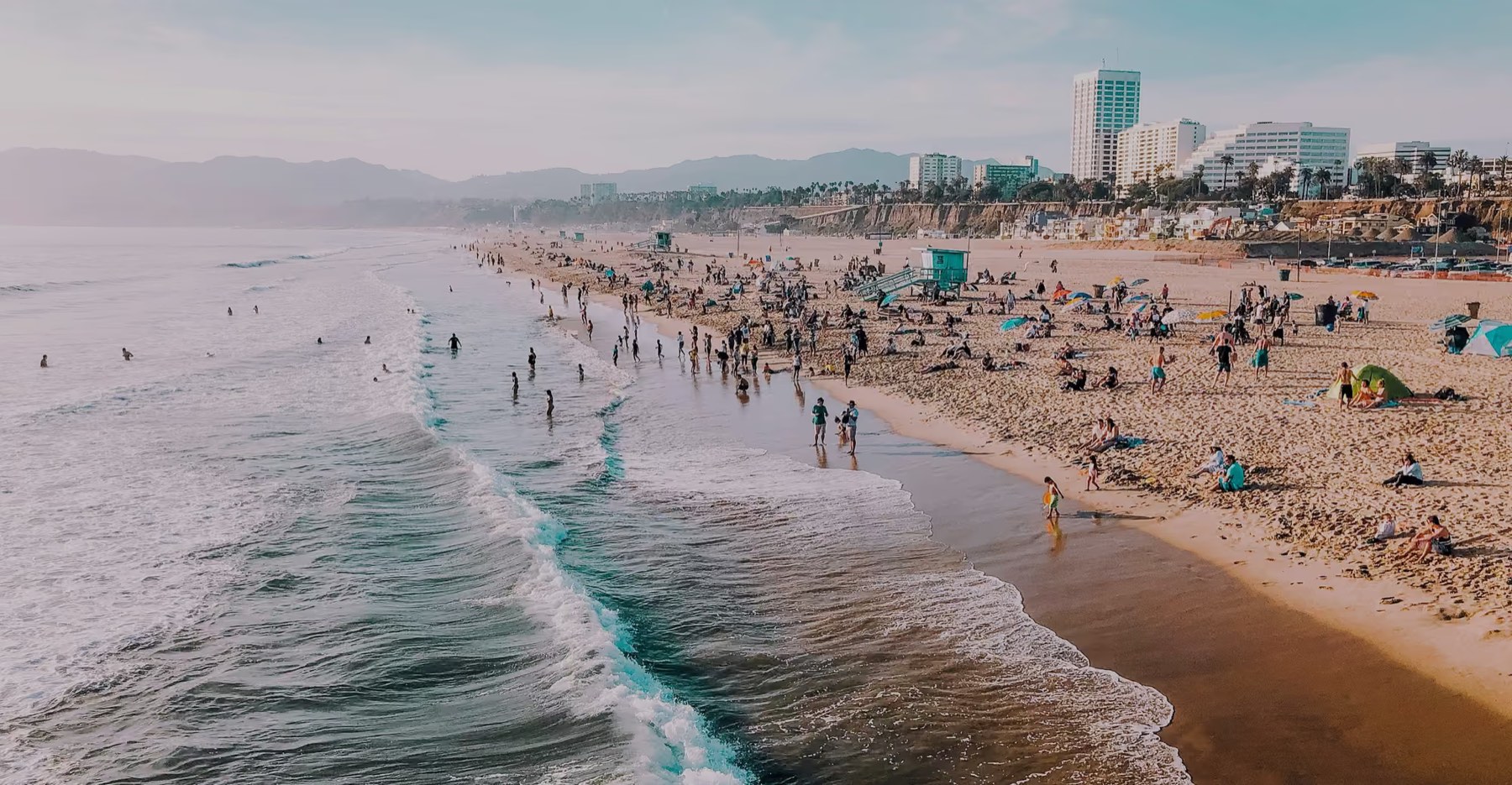 Aerial view of Santa Monica State Beach with swimmers in the surf, a lifeguard tower and the Santa Monica skyline behind the sand